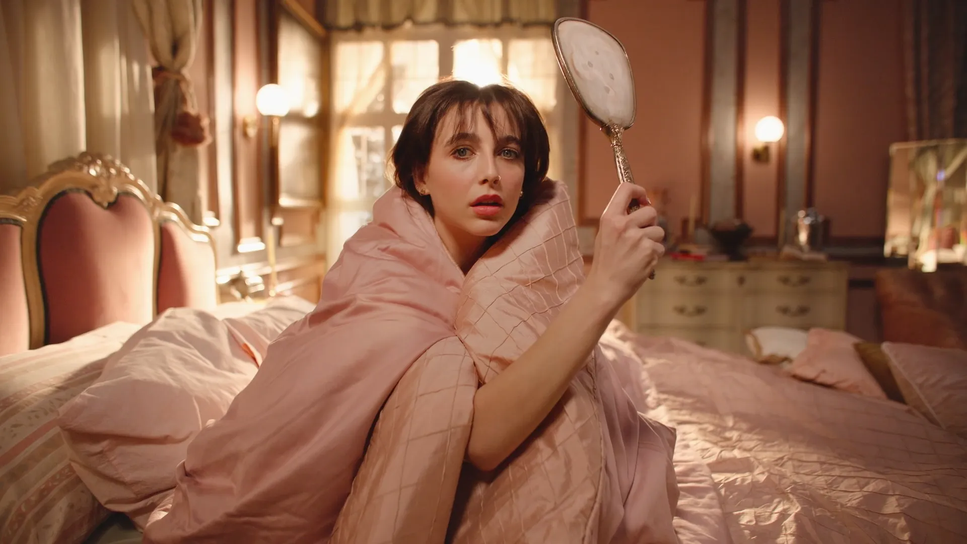Emma Chamberlain holding a hand mirror in a warmly lit room.